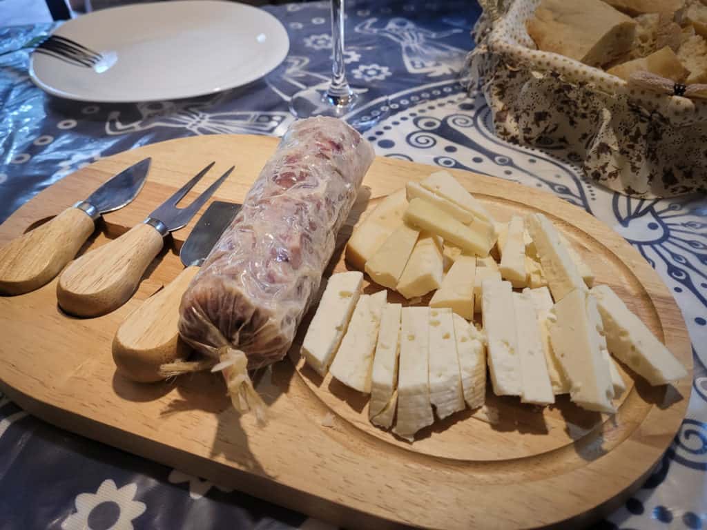 A brown plate of cheese and a log of sausage with various cheeseboard utensils in front of a bowl of bread