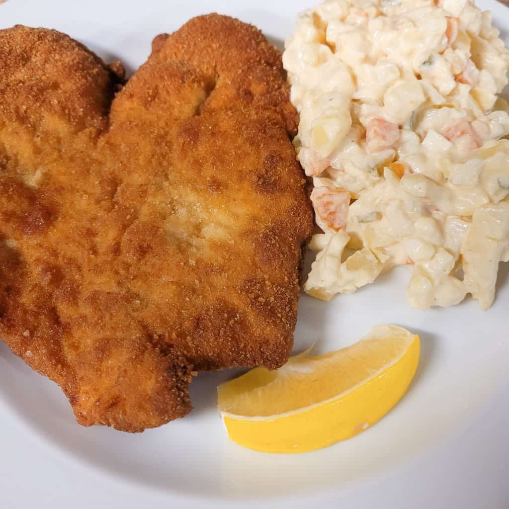 Breaded pork cutlet next to potato salad served with a slice of lemon wedge