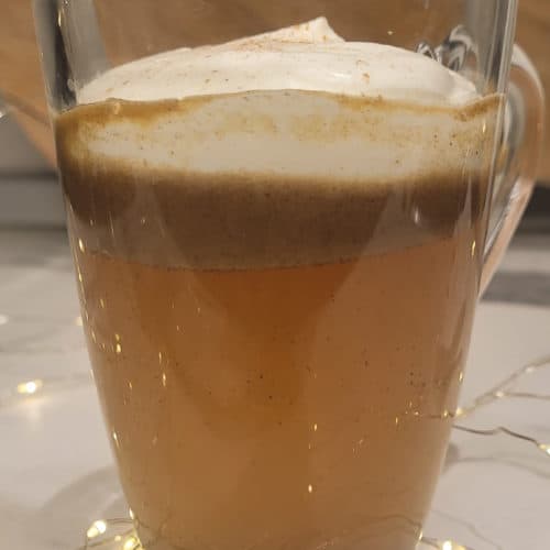 A clear mug with hot buttered rum topped with whipped cream surrounded by string lights