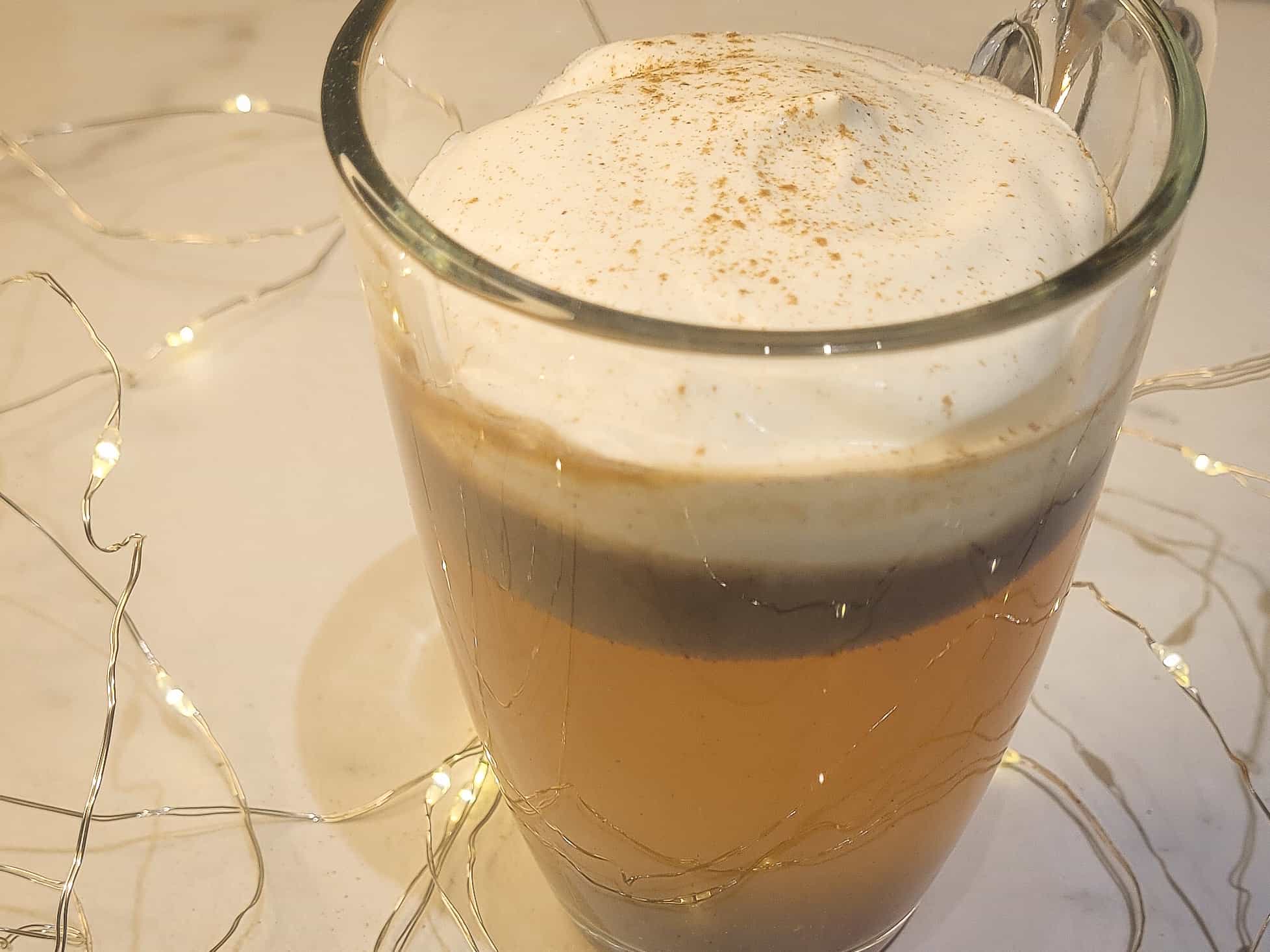 A closeup of a clear mug with hot buttered rum topped with whipped cream surrounded by string lights.
