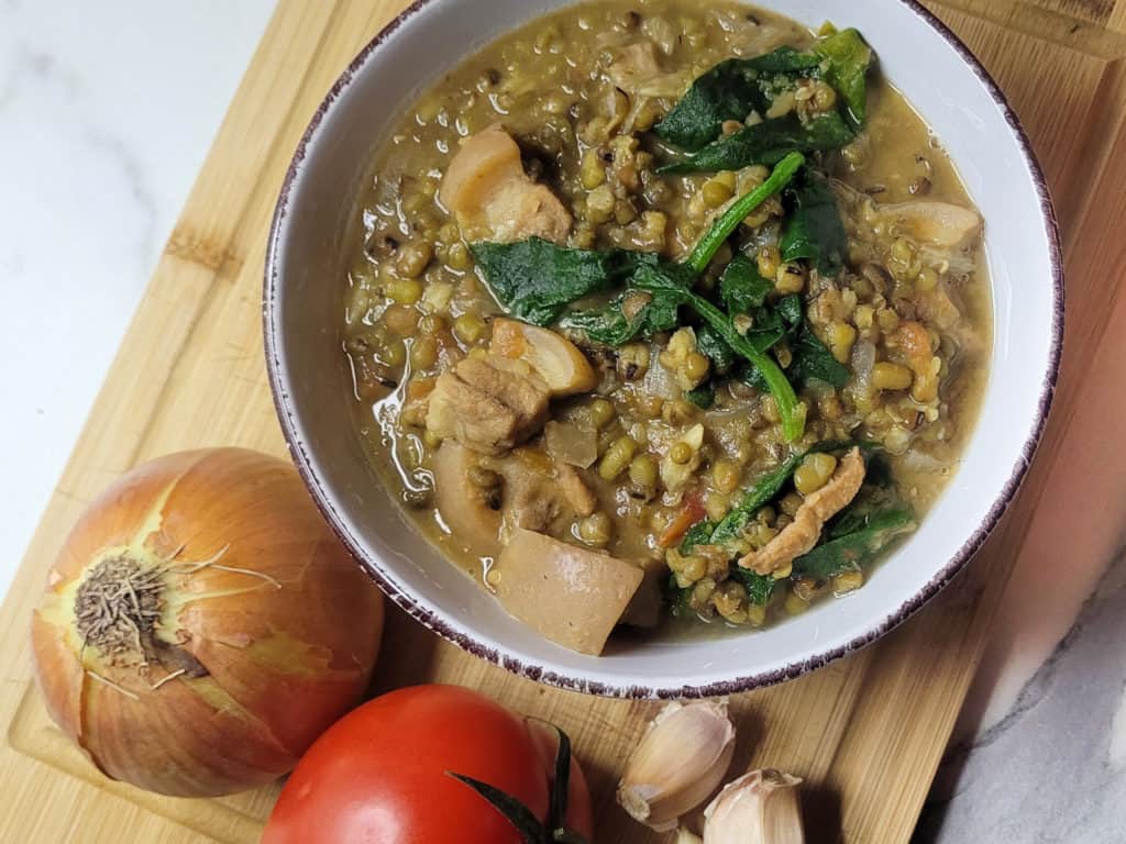 Filipino mongo in a white bowl next to an onion, tomato and some garlic cloves