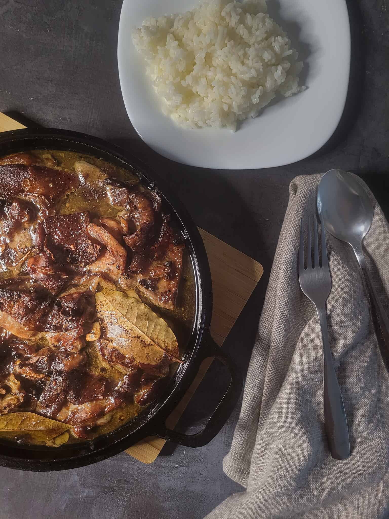 A cast iron full of chicken adobo next to rice and silverware