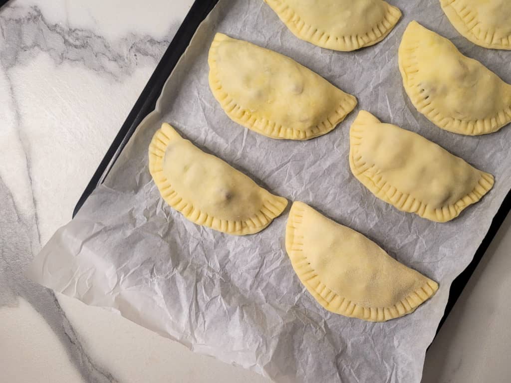 Raw empanadas on a lined baking sheet on a counter