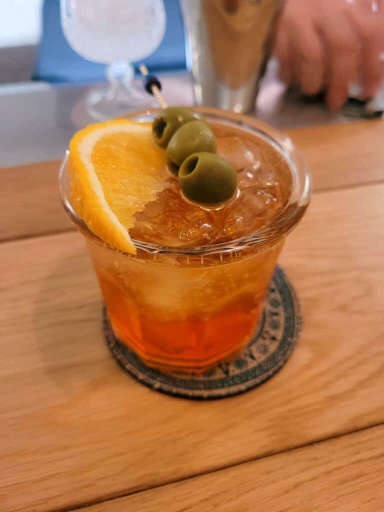 Orange cocktail in a clear glass with an orange slice and stick of olives