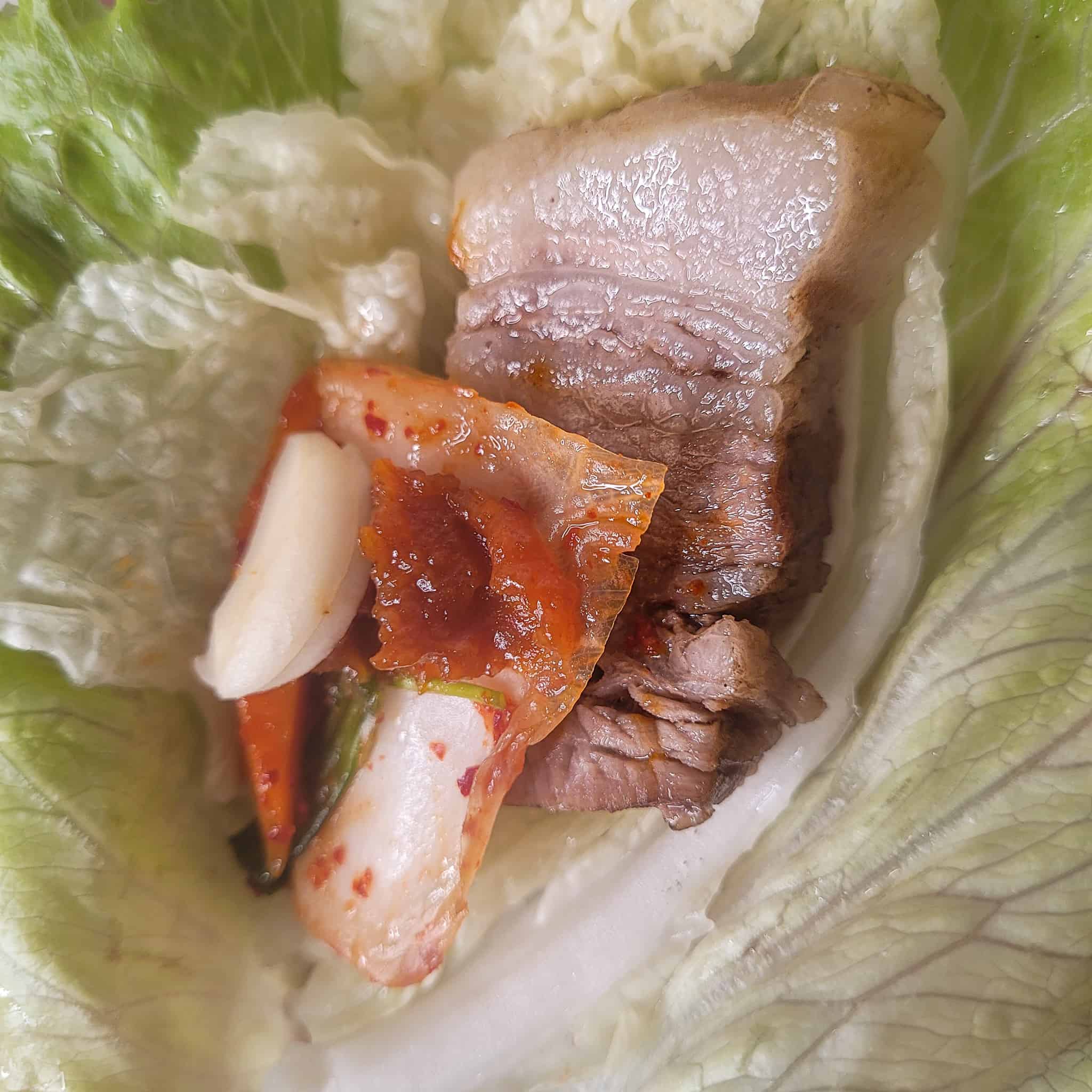 Closeup of a slice of boiled pork wrap (bossam) with kimchi on a lettuce leaf.