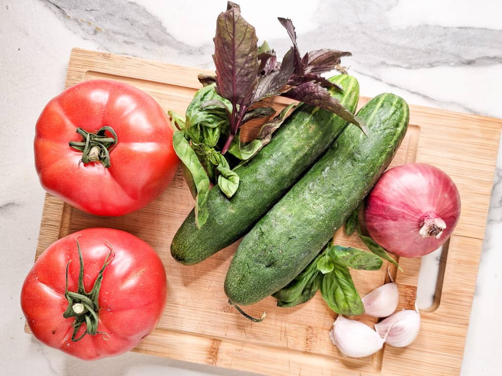 Cucumbers, basil, tomatoes, red onion, and garlic on a wooden cutting board