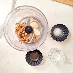Walnuts and garlic cloves in a food processor next to salt, water, and vinegar