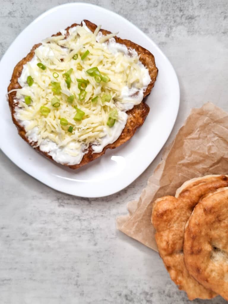 A langos or Hungarian deep fried bread with a topping of sour cream, cheese, and green onions next to a stack of langos